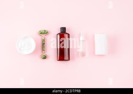 Prodotti per la cura della pelle su superficie rosa pastello. Shampoo o bottiglia cosmetic lozione, rullo di giada, asciugamano, vaso aperto crema, siero con peptidi. Naturale organico Foto Stock