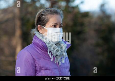 Vista laterale di bella razza mista donna di mezza età che indossa una maschera chirurgica bianca per la protezione contro Coronavirus (COVID-19). Foto Stock