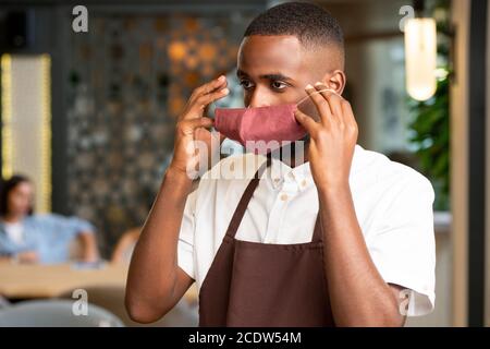 Giovane africano in uniforme di cameriere che mette maschera protettiva sul viso Foto Stock