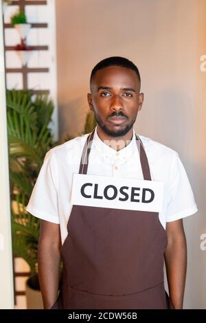 Giovane serio cameriere di etnia africana con la carta dicendo chiuso sul collo Foto Stock