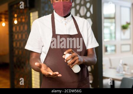 Cameriere giovane in uniforme e maschera protettiva che spruzzano il disinfettante mani Foto Stock