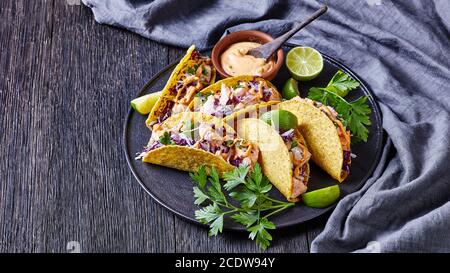 tacos di salmone con insalata di cavolo rosso con salsa piccante allo yogurt cosparsa di prezzemolo tritato finemente servito su un piatto nero su un tavolo di legno scuro, hori Foto Stock