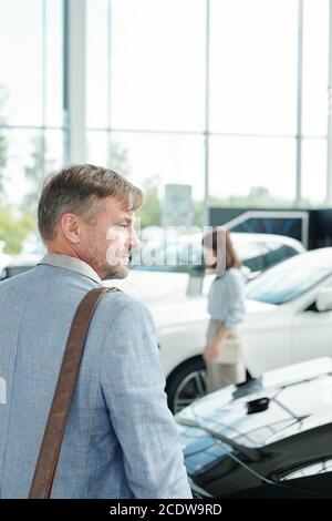 Vista posteriore di un uomo maturo in giacca blu in piedi parte anteriore di una lussuosa auto nera Foto Stock