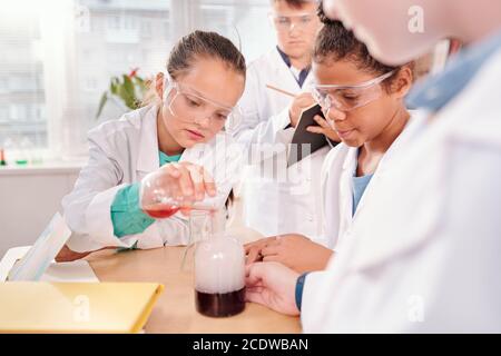 Carino studentessa mescolando due liquidi in bottiglia mentre uno di scrittura di compagni di classe Foto Stock