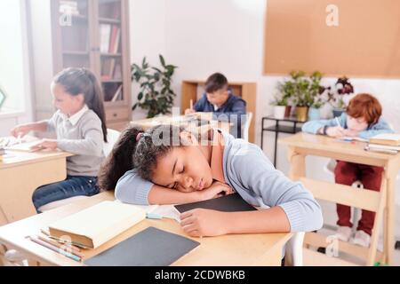 Carino scolaro stanco in casualwear che tiene la testa sulla scrivania durante il napping Foto Stock