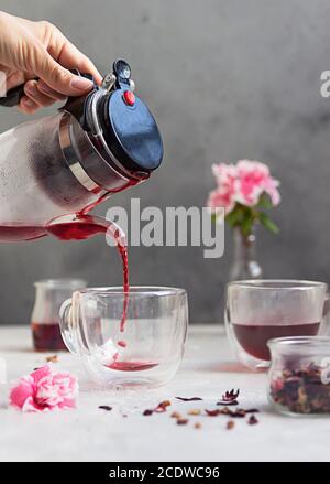 Versare il tè hibiscus caldo in una tazza di vetro e fiori rosa su uno sfondo di pietra grigio chiaro. Preparazione del tè hibiscus. Bevanda calda. Foto Stock