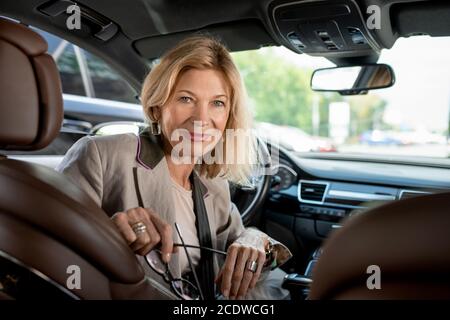 Felice donna matura seduta da Steer in auto e guardando a qualcuno sul sedile posteriore Foto Stock