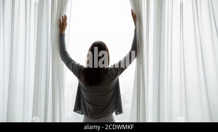 Vista posteriore giovane donna che apre le tende al mattino dopo il risveglio Foto Stock