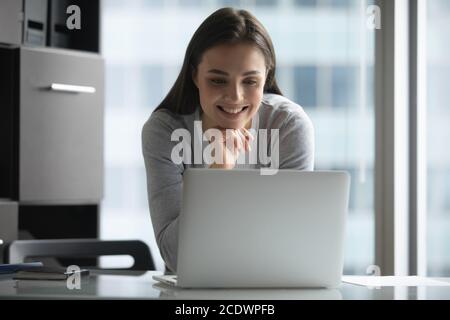 Giovane donna sorridente in piedi vicino alla scrivania, guardando lo schermo del computer portatile Foto Stock