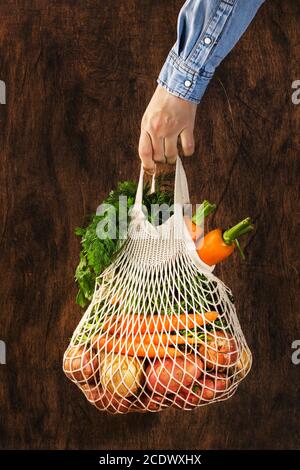 Borsa a rete con verdure ed erbe in mano femminile. Donna tenere stringa rete shopping borsa su sfondo di legno. Shopping moderno e riutilizzabile, eco-friendly lif Foto Stock