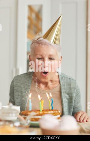 Donna anziana in cappello seduto al tavolo e soffiando le candele sulla sua torta di compleanno Foto Stock