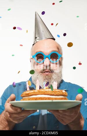 Ritratto di uomo anziano che soffia le candele nel suo compleanno torta sotto i confetti isolati su sfondo bianco Foto Stock