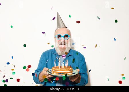 Ritratto di un uomo anziano eccitato in occhiali da sole che sorridono alla macchina fotografica mentre si è in piedi sotto i confetti e tenendo la torta isolato sopra sfondo bianco Foto Stock