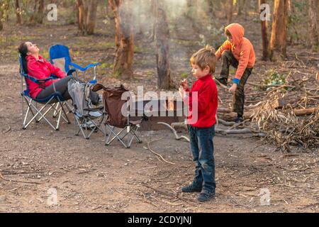 I bambini che bruciano il fuoco a Kuitpo foresta campeggio mentre loro madre che si rilassa in sedia durante le vacanze scolastiche Foto Stock