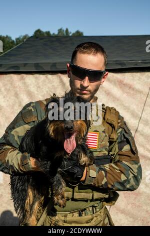 Ritratto di serio giovane soldato bearded in occhiali da sole e mimetizzazione outfit che tiene il cane carino in accampamento militare Foto Stock