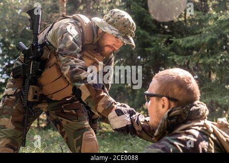 Soldato bearded in cappello e gilet che dà la mano ai feriti amico militare mentre lo raccoglie fuori terra in foresta Foto Stock