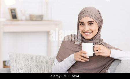 Relax domestico. Sorridente ragazza araba a Hijab rilassante sul divano con caffè Foto Stock
