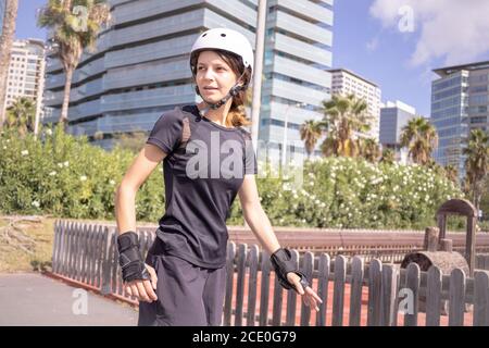 Giovane pattinatore caucasico donna nel casco bianco e nero abiti sportivi, giornata di sole, skatepark, ambiente urbano. Foto Stock