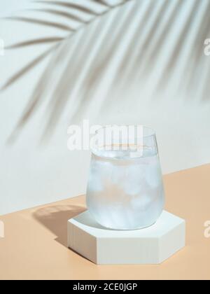 Acqua fredda con ghiaccio in vetro bicchiere sul piedistallo esagonale. Mockup per un drink in stile alla moda con foglie tropicali ombra. Moderna natura morta con vetro. Spazio di copia. Salute, bevande, concetto estivo Foto Stock
