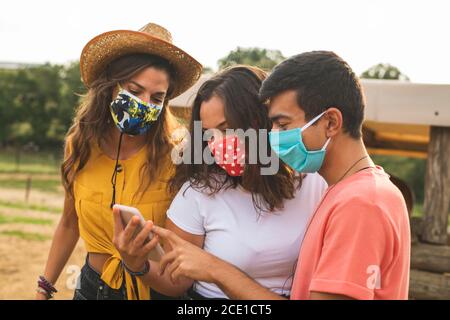 Amici divertirsi utilizzando uno smartphone in un ranch con maschere protettive. Ranch Concept Photography Foto Stock