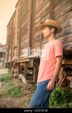 Giovane uomo con cappello Cowboy che guarda il tramonto davanti a un vagone in legno d'epoca. Ranch Concept Photography Foto Stock