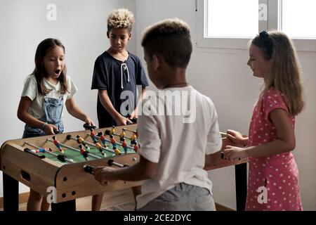 Gruppo di bambini carino multiculturale stand al coperto trascorrere il tempo di gioco con gli amici giocare a foosball insieme. Versione da tavolo degli amanti del calcio. Avendo fu Foto Stock