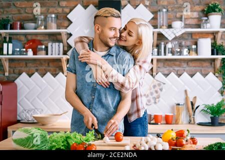 Sorridente giovane coppia che cucina insieme pasto vegetariano in cucina a casa. Donna che abbraccia l'uomo