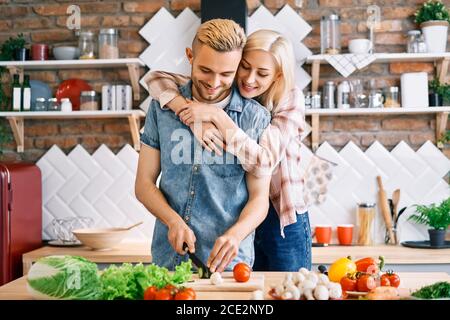 Sorridente giovane coppia che cucina insieme pasto vegetariano in cucina a casa. Donna che abbraccia l'uomo Foto Stock