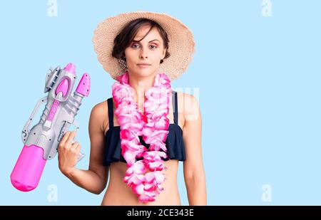 Bella giovane donna con capelli corti indossando bikini e hawaiano lei che tiene l'atteggiamento di pensiero della pistola dell'acqua e l'espressione sobria che osserva sicuro di sé Foto Stock