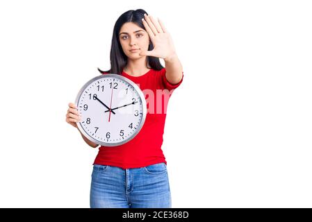 Giovane bella ragazza tenendo orologio con mano aperta facendo segno di stop con espressione seria e fiduciosa, gesto di difesa Foto Stock