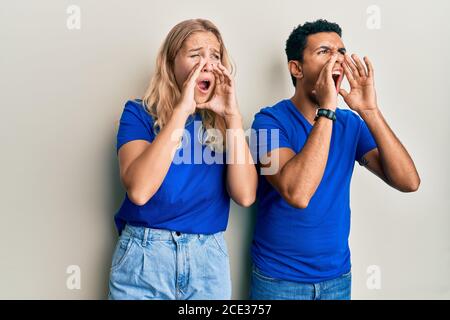 Giovane coppia interracial che indossa abiti casual gridando arrabbiato fuori forte con le mani sulla bocca Foto Stock