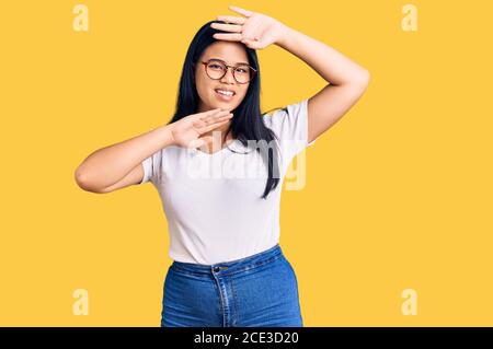 Giovane bella ragazza asiatica indossando abiti casual e bicchieri sorridente allegro giocando a peek un boo con le mani che mostrano il volto. Sorpreso ed uscito Foto Stock