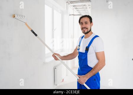 Colpo medio, uomo di lavoro in abiti da lavoro blu dipinge parete in appartamento Foto Stock