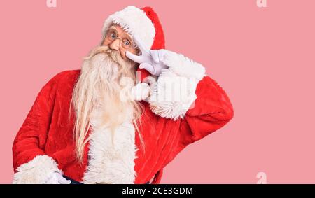 Vecchio uomo anziano con capelli grigi e barba lunga che indossa il costume tradizionale di babbo natale facendo simbolo di pace con le dita sul viso, sorridendo allegro showi Foto Stock