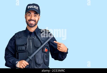 Giovane uomo ispanico che indossa uniforme polizia tenendo baton sorridente felice puntare con la mano e il dito Foto Stock