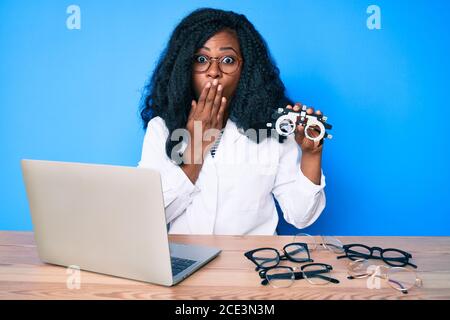 Bella donna africana che tiene occhiali di optometria che copre la bocca con la mano, scioccata e impaurita per errore. Sorpresa espressione Foto Stock