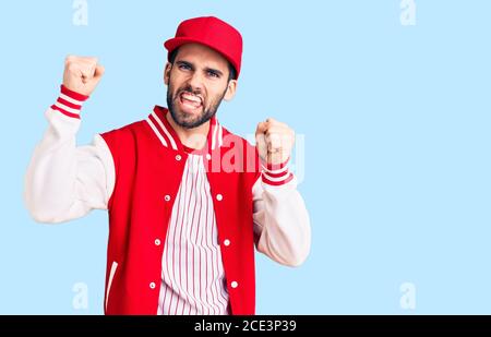 Giovane bell'uomo con barba che indossa giacca da baseball e berretto urlando orgoglioso, celebrando la vittoria e il successo molto eccitato con le braccia sollevate Foto Stock