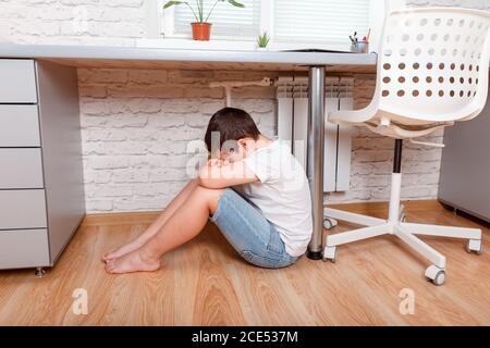 Ragazzo sconvolto. Il ragazzo adolescente si siede da solo sotto la scrivania, coprendo il viso Foto Stock