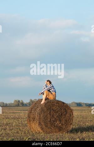 Romantica donna bruna in una camicia plaid e pantaloni seduta su un fiammifero rotondo in un campo incredibile, sullo sfondo della foresta. Colori caldi dei soli Foto Stock