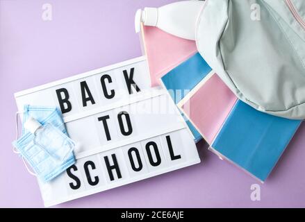 Scatola luminosa con testo DI RITORNO A SCUOLA maschera protettiva viso e igienizzatore. Nuovo normale. Libri che cadono dallo zaino. Distanza sociale Foto Stock