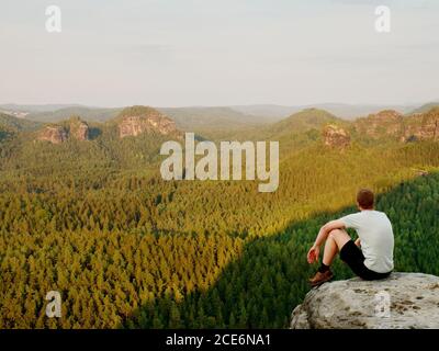 Momento di solitudine. Uomo in camicia sedersi sulla scogliera di roccia e guardare nella valle della foresta. Foto Stock