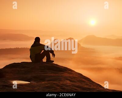 Paesaggio primaverile. Escursionista in nero sulla cima rocciosa. Meraviglioso break in montagna, forte nebbia arancione in profonda valle. Uomo siede o Foto Stock