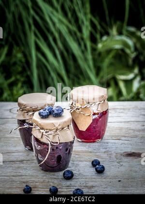 Tre vasetti di marmellata fatta in casa da more e lamponi su tavola di legno d'annata nel giardino. Primo piano con spazio per la copia. Shot verticale. Foto Stock