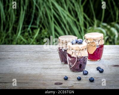 Tre vasetti di marmellata fatta in casa da more e lamponi su tavola di legno d'annata nel giardino. Primo piano con spazio per la copia. Foto Stock