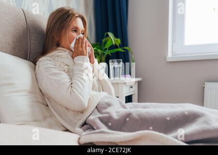 La giovane donna soffre di influenza a casa. La femmina soffia il naso che tiene il fazzoletto del tessuto, avendo sintomi di sinusite cronica, allergia stagionale, Foto Stock