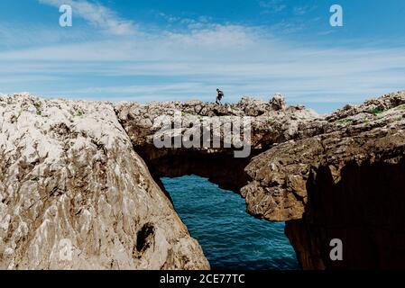 Vista laterale dell'anonimo viaggiatore maschile esplorando scogliere ruvide con superficie irregolare sopra acqua increspata oceano sotto cielo spettacolare con nuvole durante il vca Foto Stock