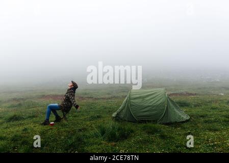 Full length soddisfare femmina viaggiatore in jeans e caldo khaki giacca seduta su sedia pieghevole vicino tenda da campeggio con tazza di bevanda calda durante il campeggio in alture con occhi chiusi Foto Stock