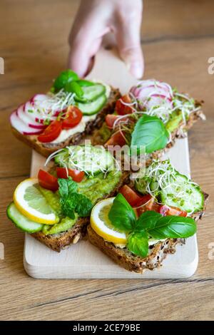 Coltivare femmina con deliziosi toast con cetrioli e pomodori guarniti con limone fresco e ciuffi di basilico e posto sopra tagliere di legno Foto Stock