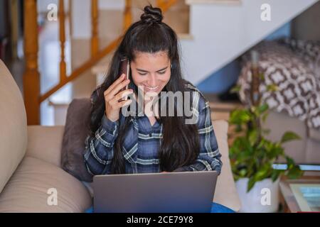 Giovane ragazza allegra freelancer in abito casual seduto sul divano con il computer portatile in ginocchio e mentre parli con il telefono cellulare lavorare a un progetto remoto a casa Foto Stock