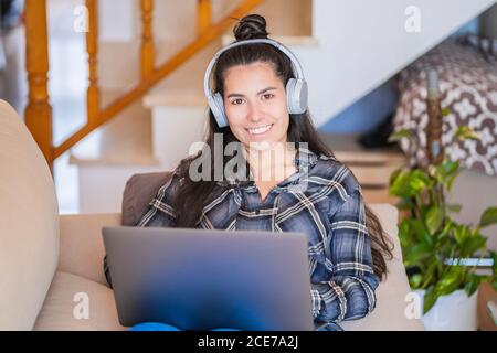 Giovane studentessa di abbigliamento casual con cuffie che guardano video lezione sul computer portatile mentre si siede sul divano in un appartamento moderno Foto Stock